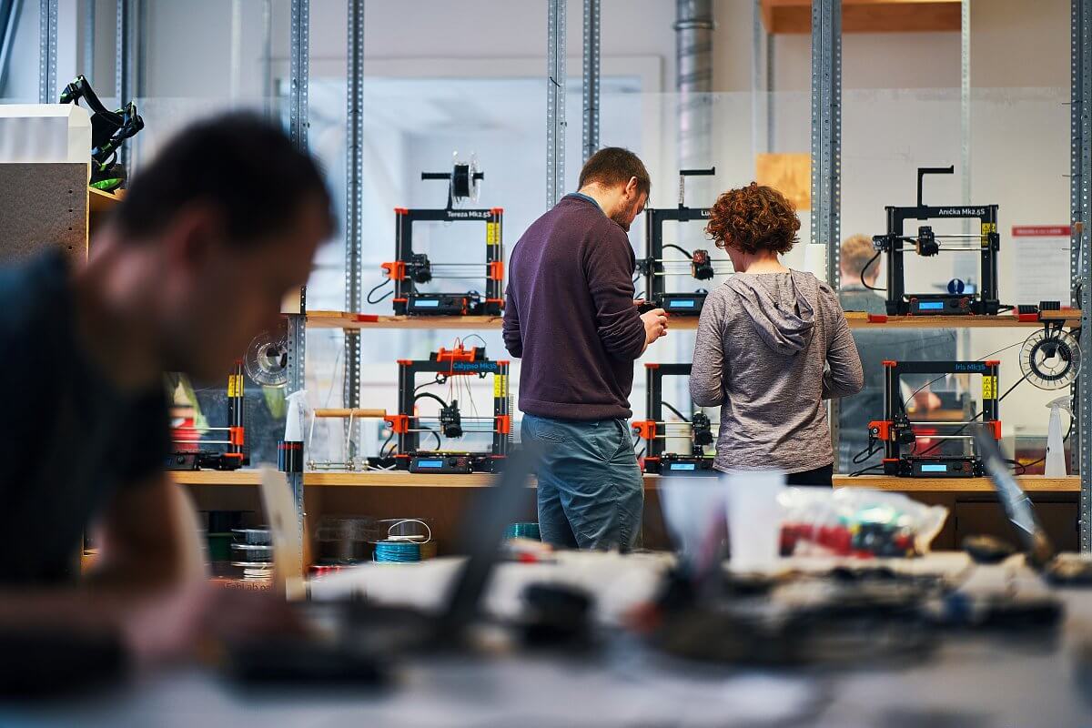 Dva studenti prohlížející výrobek před řadou 3D tiskáren v univerzitní laboratoři.